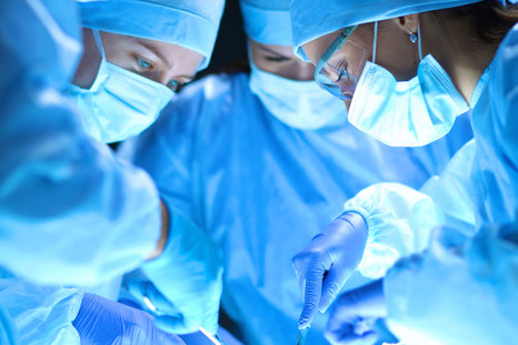 Healthcare professionals in blue surgical gowns, masks, gloves, and caps are leaning over an operating table.