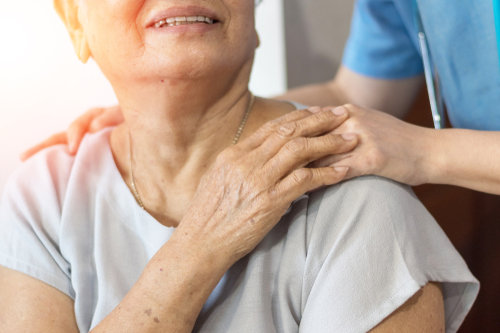 Someone is resting a hand on an older woman's shoulder. The older woman has her hand on top of  the person's hand and is looking up at them.